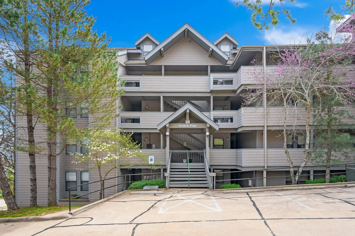 Exterior view of the building’s entrance, framed by mature trees, with balconies that overlook the landscape. The scene is inviting and suggests a quiet retreat above the trees, possibly with access to lake views.