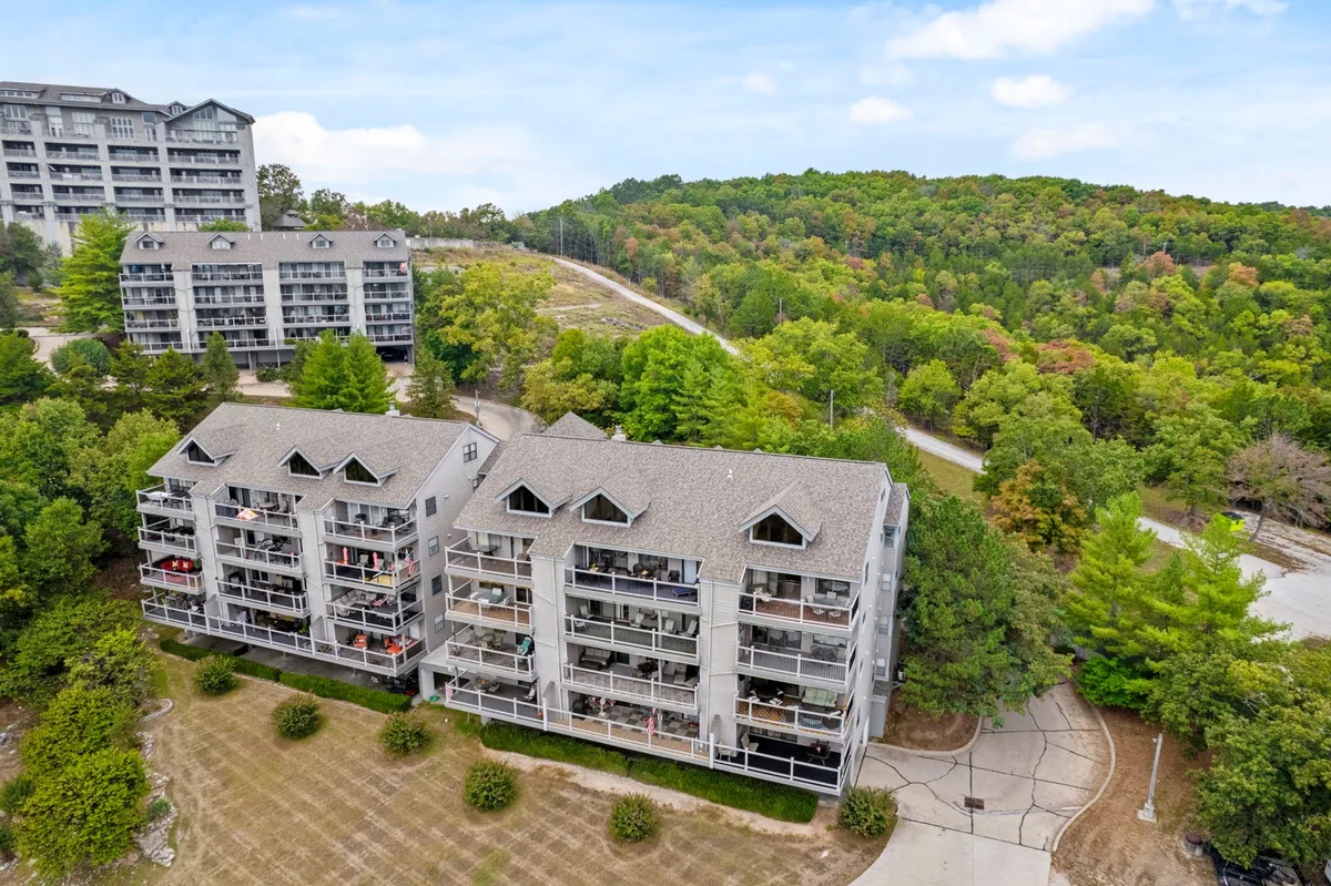 A peaceful exterior view of three-story condo buildings nestled among mature trees, with a winding pathway leading through the greenery. Residents can be seen relaxing on balconies and strolling along the path, enjoying the connection to nature.