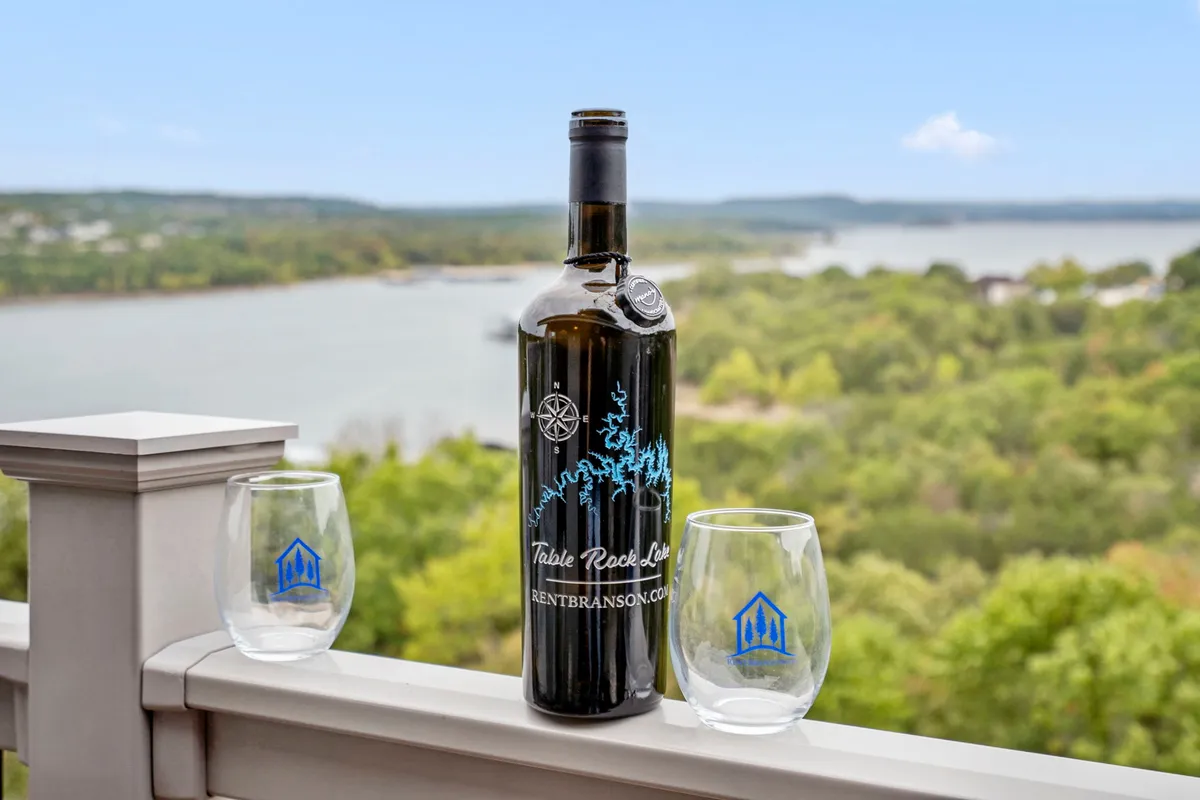 A serene exterior shot from the private deck features a bottle of wine and two glasses overlooking a scenic lake view, evoking leisurely moments like sipping coffee or wine while taking in the landscape.