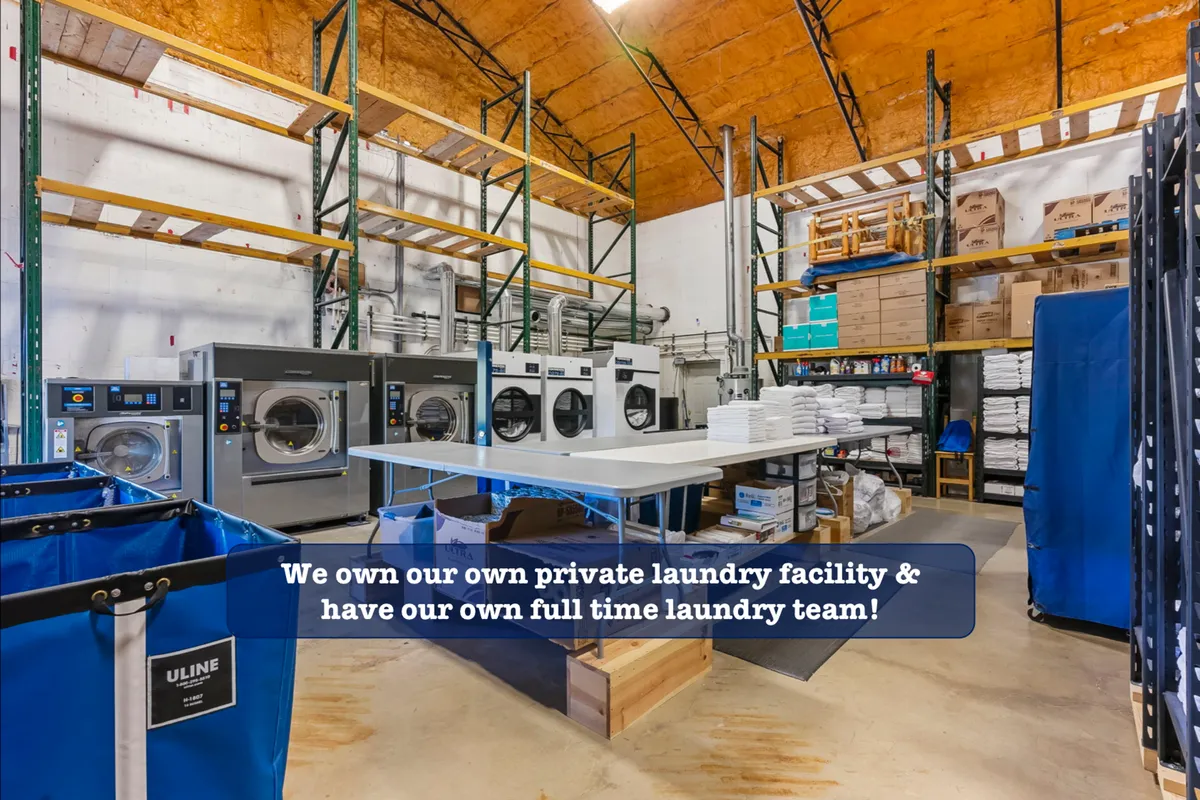 A professional laundry facility with industrial washing machines, folding tables, and storage shelves stacked with linens. The space is bright, organized, and efficient, communicating high standards of cleanliness.