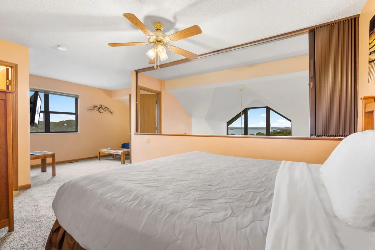 A bright bedroom with crisp white bedding on a large bed, a ceiling fan overhead, and expansive windows letting in streams of natural light. The windows offer a partial view of the outdoors, creating an open and airy atmosphere.