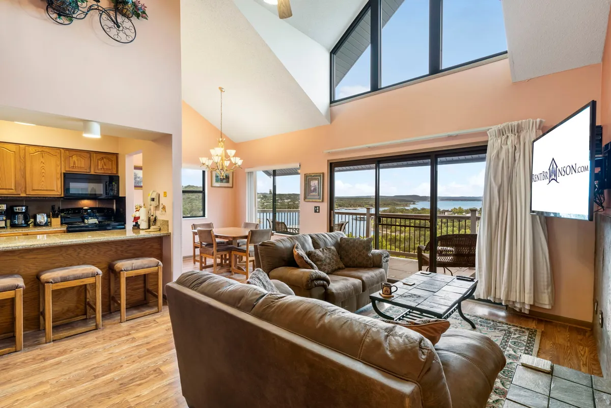 This image offers a wide view of the living, dining, and kitchen areas, with large windows framing a stunning view of the outdoors. Plush, comfortable seating and a dining table underscore the room’s versatility for family gatherings, relaxation, and enjoying the scenery.